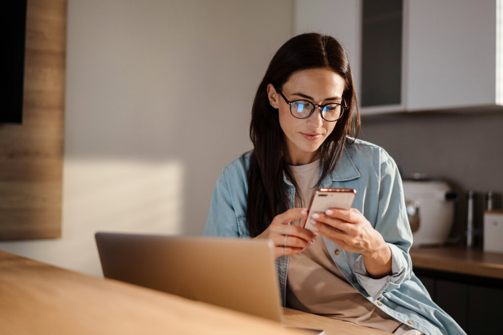 Woman with glasses using smartphone while working on her laptop