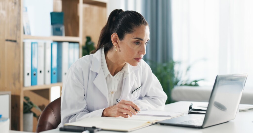 A woman in a lab coat sits at a desk, focused on internet marketing strategies displayed on her laptop among papers and a notebook.